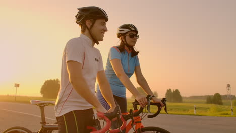A-man-and-a-woman-in-helmets-with-bicycles-stand-and-talk-at-sunset.-Rest-after-a-bike-ride-on-the-highway.-Track-bikes.-Couple-sports